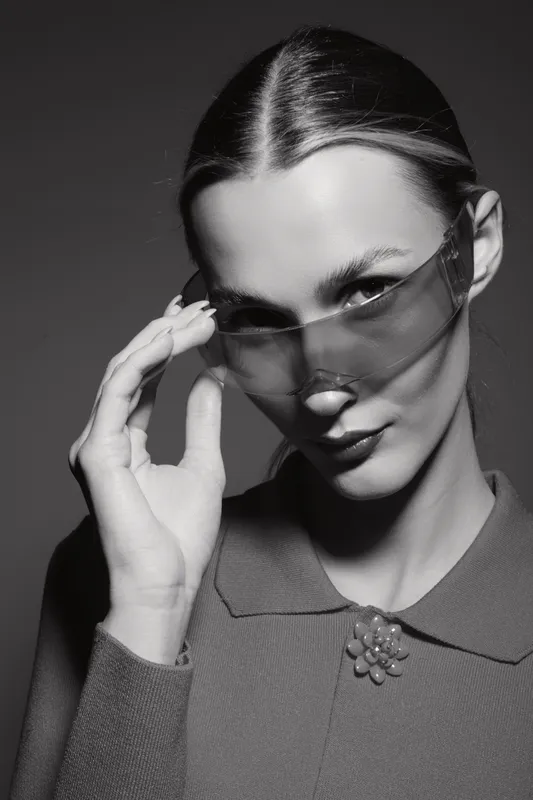 Portrait of a young white woman with slicked hair wearing sunglasses and a textured top in studio light.