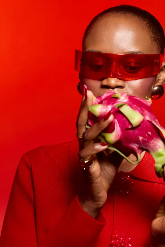 Stylish black woman with dark skin wearing red sunglasses and red outfit holds a dragon fruit close to her face against a vibrant red background. Concept of fashion and exotic fruit.