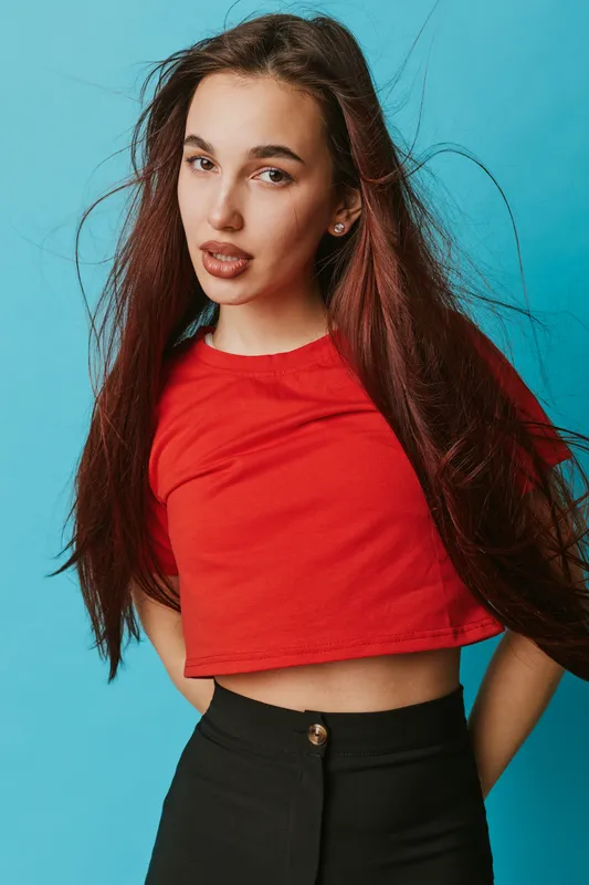 Young woman with long dark hair wearing red crop top and black pants posing against blue background. Portrait of confident female model with natural makeup and casual style indoors.