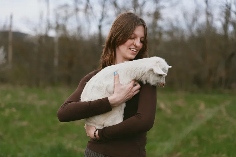 Young woman with brown hair gently holds a white baby goat outdoors in a natural green field during a calm spring day.