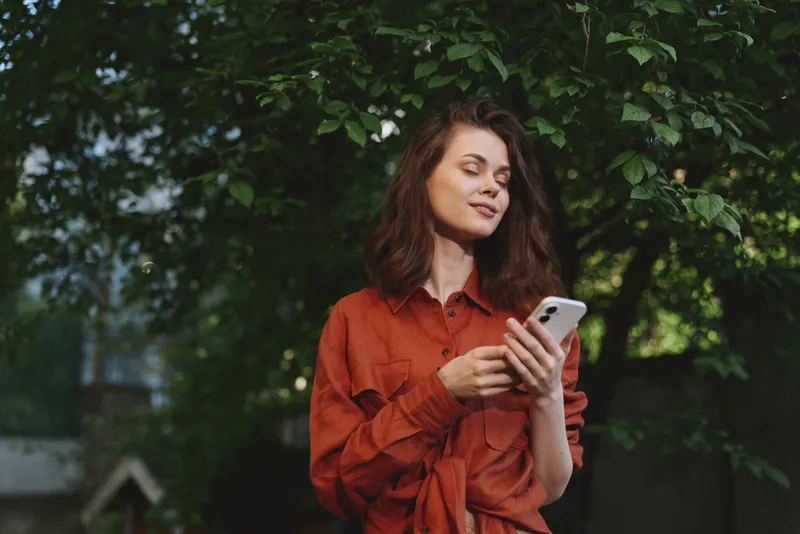 Young woman with shoulder length brown hair stands outdoors, looking at her smartphone with a gentle smile, wearing a rust red shirt amid green foliage