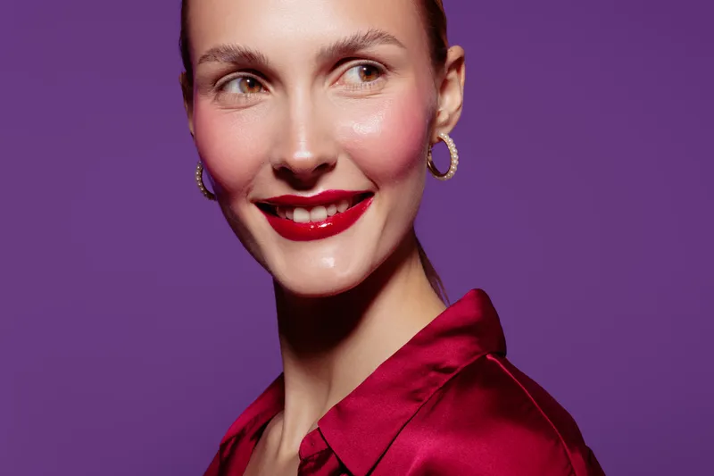 Smiling woman with red lipstick and hoop earrings looks sideways on purple background. She wears a red satin blouse. Concept of beauty, fashion, and confident female portrait.