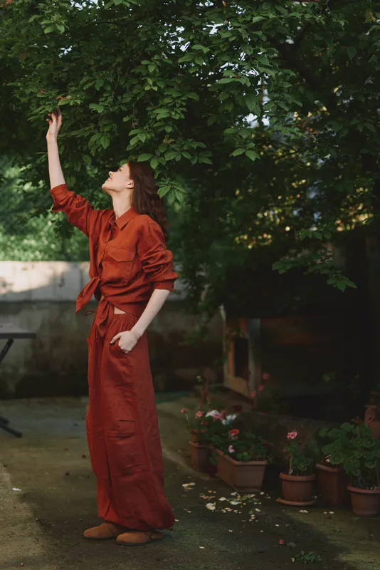 A woman in a red dress raises her arm toward the sky in a sunlit garden, conveying a moment of freedom, confidence, and playful movement