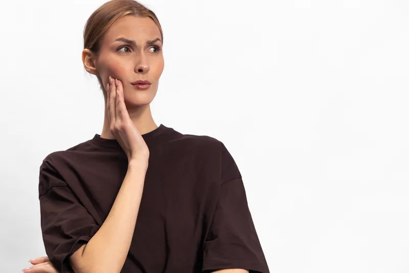 Concerned young woman in dark shirt touches cheek with hand, looks away with thoughtful expression. Isolated on white background. Concept of pain, discomfort, and contemplation.
