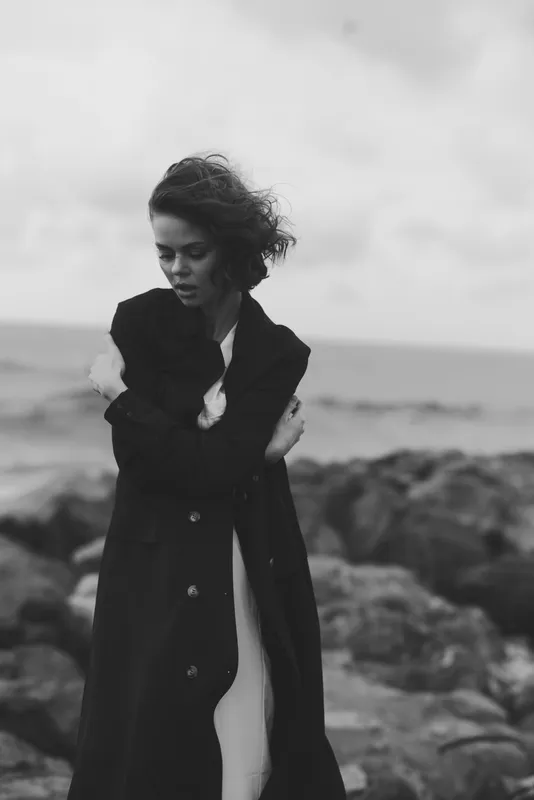 Moody black and white scene of a woman in a long coat on a windy rocky coast, hair swept by wind, dramatic seaside atmosphere, expressive fashion mood and solitary strength