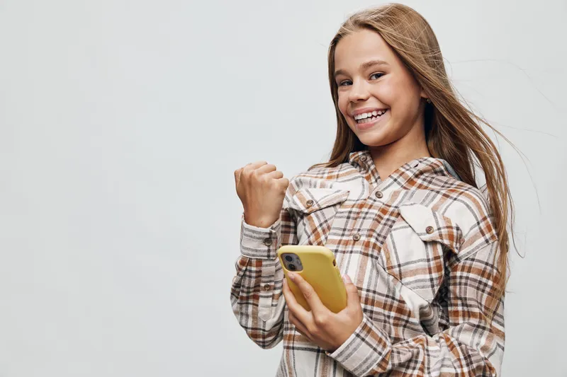 Teenage girl smiling and looking at camera holding smartphone with yellow case in one hand and raised fist celebrating success or victory on light background.