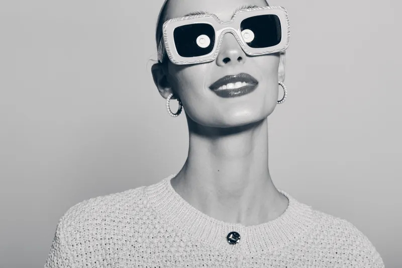 Stylish white woman with pale skin wearing oversized white sunglasses and hoop earrings looks confident in black and white studio portrait.