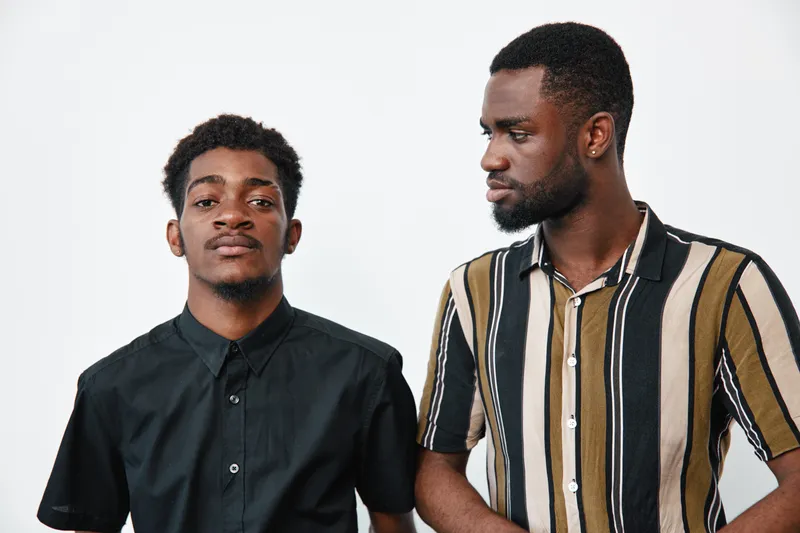 Two african american men with dark skin standing together in casual clothing on white background. One man looks at camera while the other looks at him with serious expression.