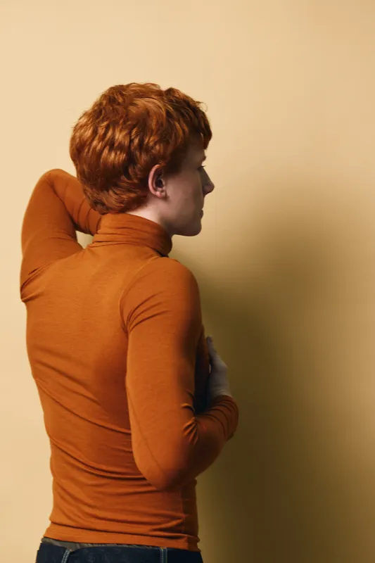 Young redhead man in orange turtleneck sweater posing sideways with hand behind head on beige background in studio. Casual style, thoughtful mood and profile portrait.