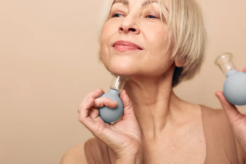 Mature woman with short blonde hair uses facial massage tool on neck, smiling softly with closed eyes. Concept for skincare, anti aging, wellness, beauty, and self care at home.