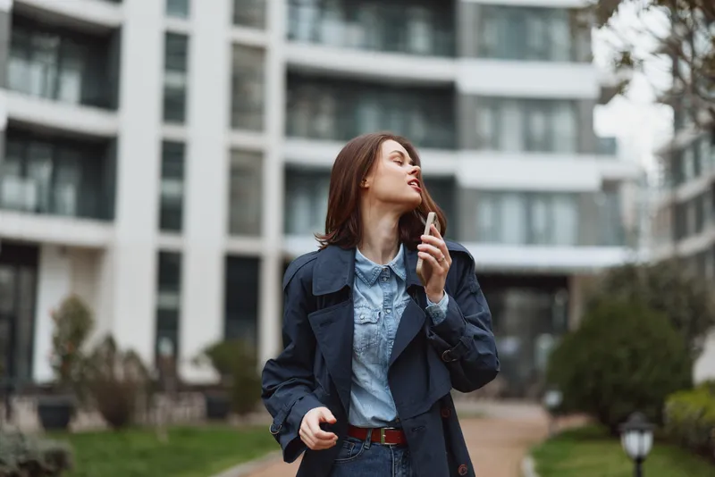 A stylish woman in a denim jacket and blue shirt walks along an urban pathway with modern buildings in the background, expressing confident street style and quiet contemplation.