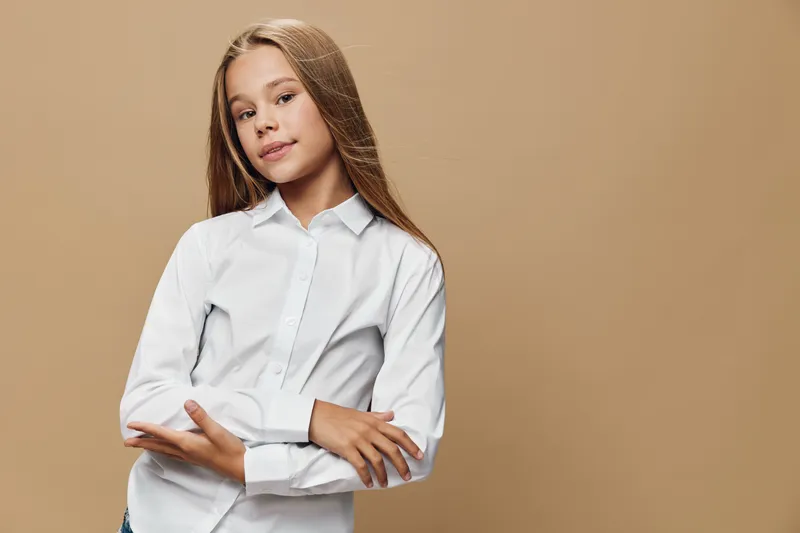 Girl with long hair wearing white shirt standing with crossed arms looking confident and calm on beige background. Portrait, youth, fashion, casual style and expression.
