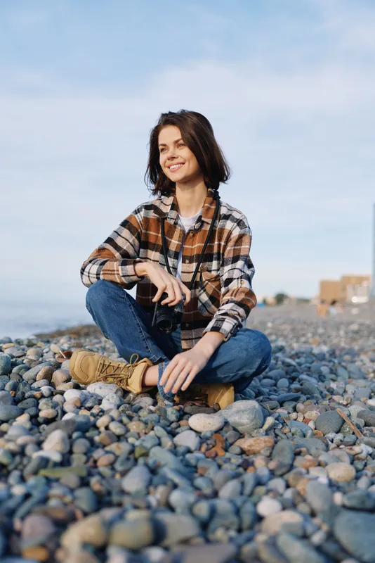 Woman sits on a pebble beach in casual plaid shirt and jeans with boots, smiling, enjoying outdoor relaxation by the shore and the open sky above