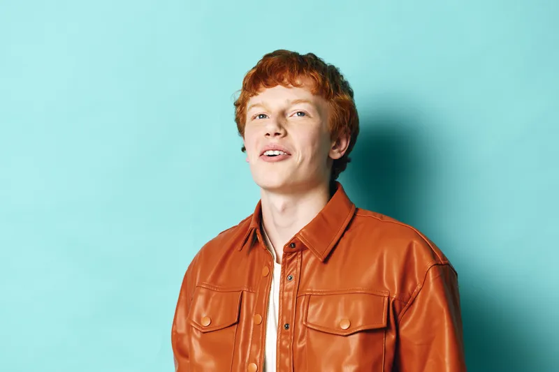 Young man with red hair wearing brown leather jacket standing against blue background posing confidently with slight smile and relaxed posture in studio lighting.