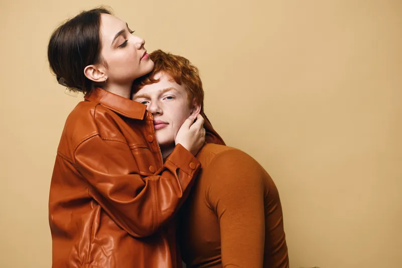 Two women in brown clothing embrace in warm hug with eyes closed on beige background. Intimate moment, affection and comfort captured in studio with casual outfits.