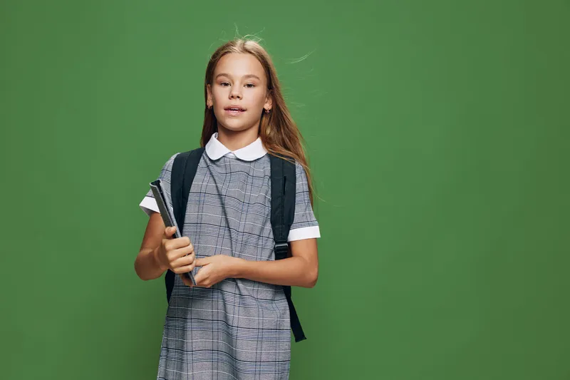 Girl with backpack and book standing against green background wearing school uniform for education and learning concept with confident expression in studio.