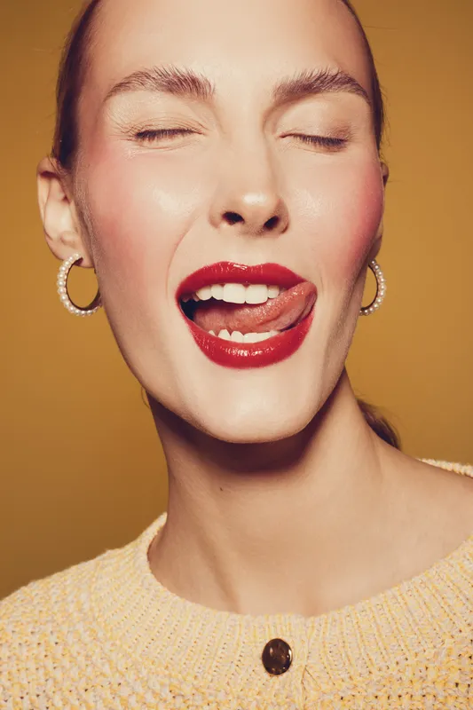 Close-up of a young woman with red lipstick winking and sticking out her tongue. She wears hoop earrings and a yellow sweater against a warm brown background. Playful and confident mood.