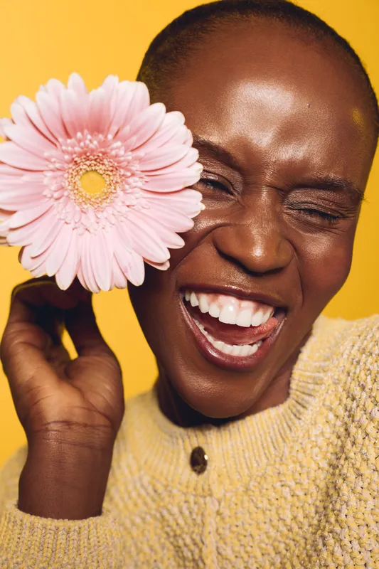 Happy African woman with dark skin laughing and holding a pink flower near her face. Yellow background and cozy sweater create warm joyful mood. Concept of beauty, happiness, and nature.