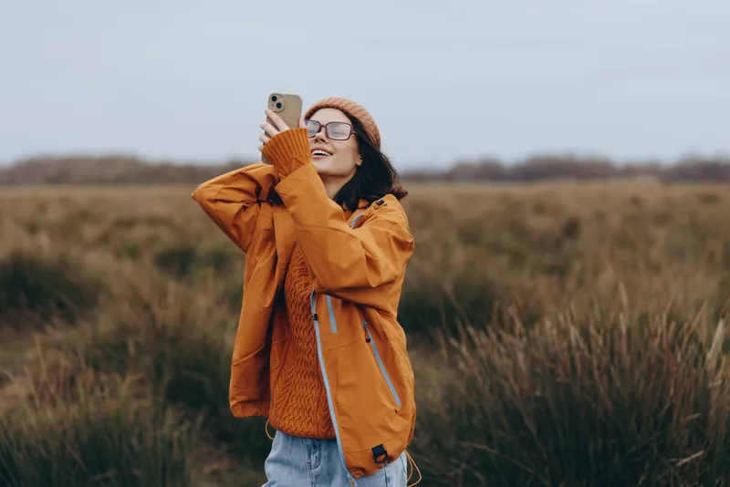 Woman in outdoor field lifestyle during autumn, wearing jacket and scarf, standing in nature countryside, smiling while holding a small dog companion in grass, warm daylight, calm mood
