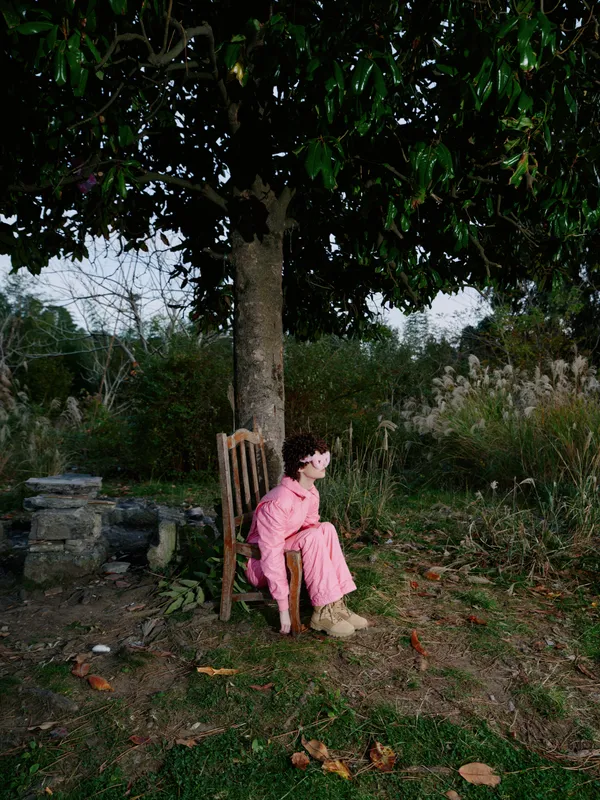 Child girl sitting on a chair beneath a tree in a garden, outdoors in pink, a peaceful rustic moment of solitude, wonder and imagination in a quiet afternoon.