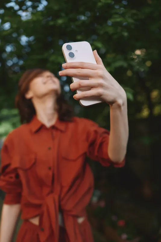 A woman in a red shirt takes a selfie in a leafy park, capturing a casual lifestyle moment. Bright natural light enhances a candid outdoor portrait