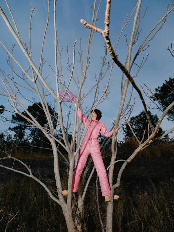 Child climbing a leafless tree in pink jumpsuit, reaching for a kite against a clear blue sky, outdoor play and balance in a open field during windy day