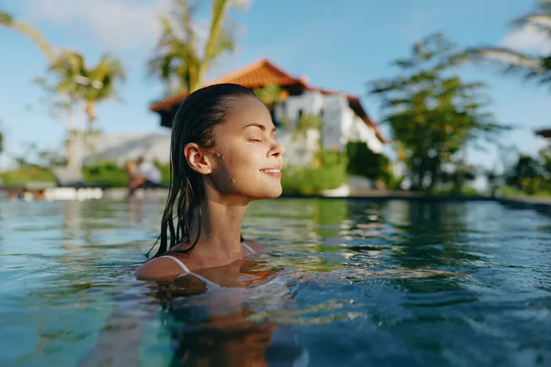 A woman relaxes in a calm pool with eyes closed, a peaceful smile, tropical resort backdrop, bright sky. This lifestyle concept highlights leisure, wellness, and mindful escape in a sunny retreat.