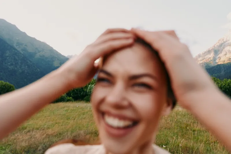 Young woman with short hair smiles joyfully while holding her head outdoors in a grassy mountain landscape under soft natural light.