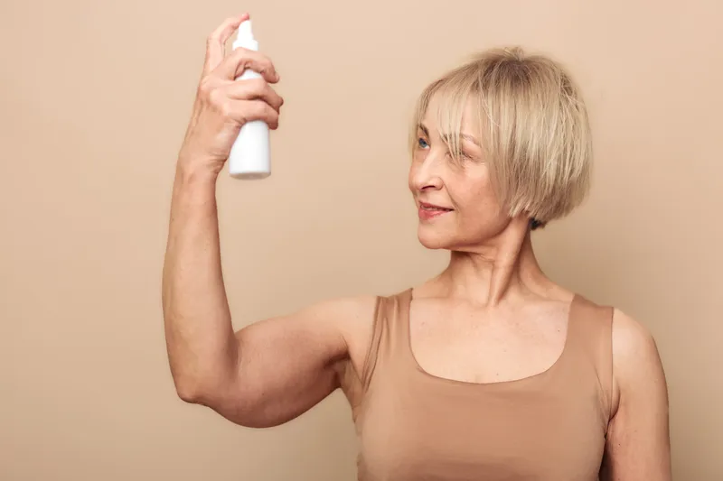 Mature woman with short blonde hair sprays facial mist on her face, wearing a beige tank top. Concept of skincare, hydration, and self-care routine for aging skin.