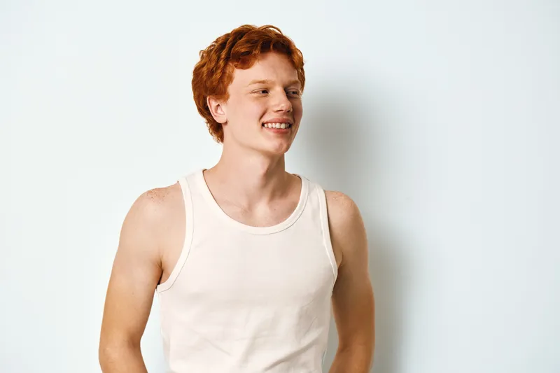 Young man with red hair wearing white tank top standing against white wall with smile, looking aside in casual clothing with bright and clean studio background, portrait from waist up.