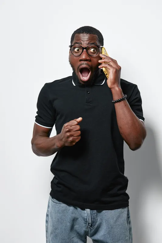 African American man with dark skin and glasses wearing black shirt talking on phone with surprised expression and clenched fist on white background.