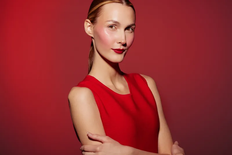 Young woman in red sleeveless dress poses confidently against red background. She has light skin, red lipstick, and a sleek hairstyle. Concept of fashion, beauty, and elegance.