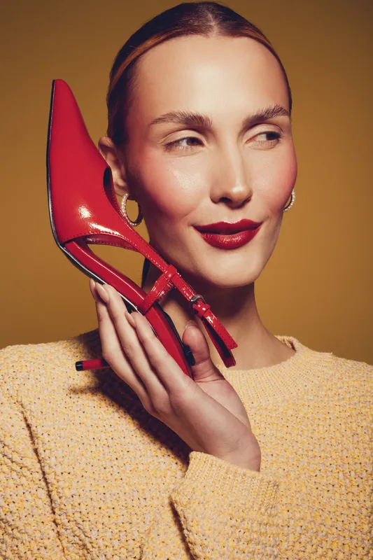 Young woman with red lipstick holds red high heel shoe near face, wearing yellow sweater. Studio portrait on yellow background. Concept of fashion, style, and beauty.