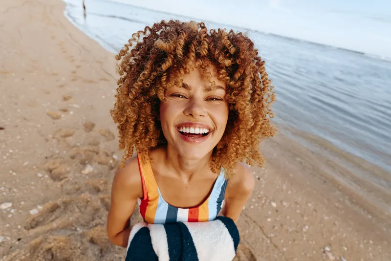 A joyful woman with curly hair on a sunny beach, smiling at the camera. Warm light, relaxed mood, and carefree summer vibes in a natural portrait.