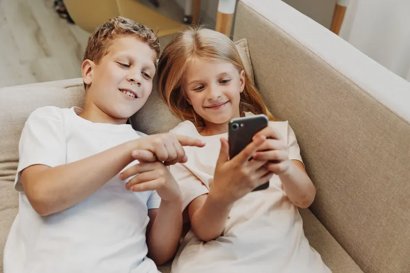 Two children lying on a couch looking at a smartphone, smiling and interacting together. Concept of family, technology, leisure, childhood, and digital communication.