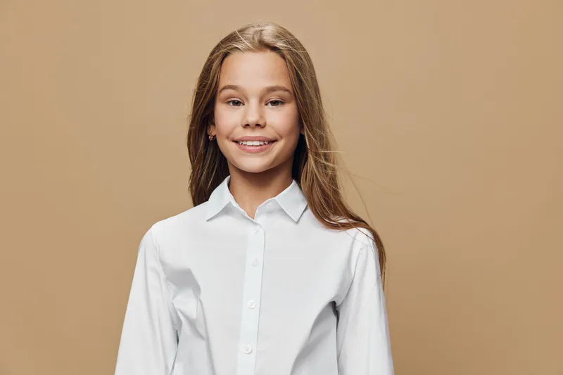 Smiling girl wearing white shirt standing against beige background with long hair and natural expression for portrait and lifestyle.