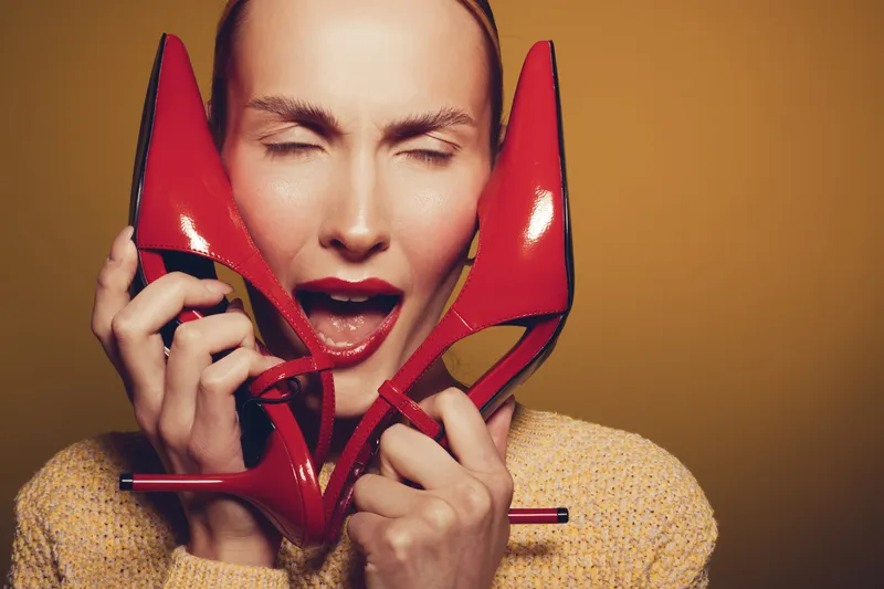Young woman with closed eyes holds red high heels near face, expressing excitement and joy. Warm lighting highlights her skin and beige sweater. Concept of fashion and happiness.