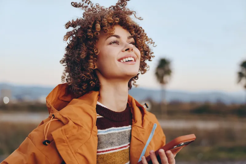 Woman with curly hair smiles outdoors at sunset, orange jacket and sweater, holding smartphone, cheerful lifestyle portrait, shallow depth of field, open field background