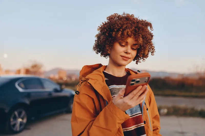 woman curly hair jacket smartphone phone outdoors lifestyle street communication, young adult holding smartphone roadside, golden hour light, casual fashion, relaxed smile, urban soft blur