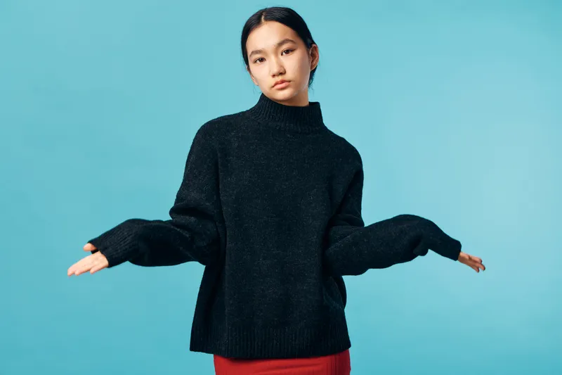 Young woman in black sweater standing with raised hands and confused expression on blue background. Casual fashion, isolated, posing and emotion of uncertainty or doubt in studio.