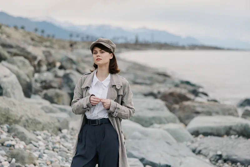 Young woman in vintage outfit stands on rocky beach with calm sea and mountains in background. She looks thoughtful, wearing a hat, coat, and high-waisted pants. Outdoor fashion concept.