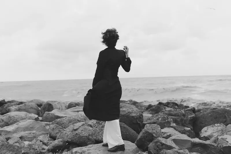Woman walks along a rocky coastline, wind catching her coat as waves crash beyond the rocks. Monochrome scene conveys solitude and resilience against the vast ocean