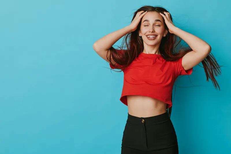 Young woman with long hair wearing red crop top and black pants posing with eyes closed and hands on head against blue background smiling with joy and confidence.