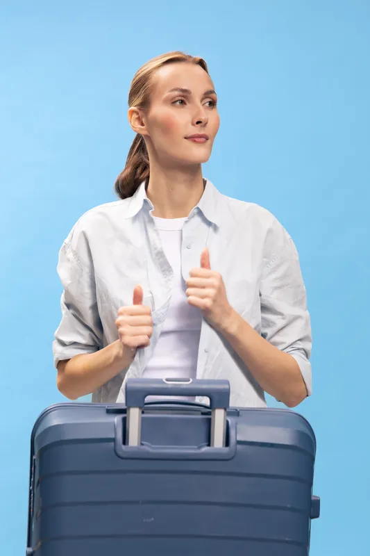 Young woman with suitcase shows thumbs up gesture, wearing casual white shirt and standing on blue background. Concept of travel, vacation, and positive mood.