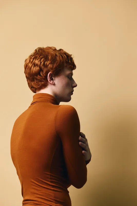 Young redhead woman in brown turtleneck standing sideways with arms crossed against beige background, portrait with thoughtful and calm expression.