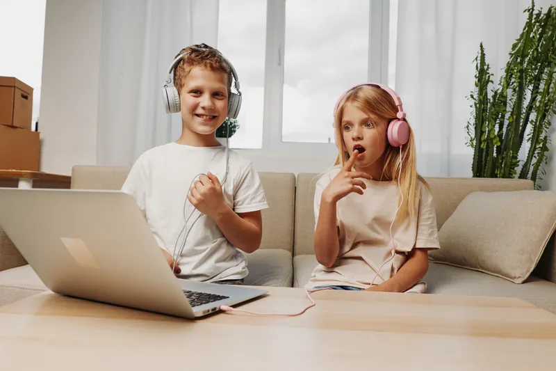 Two children wearing headphones sit on a couch with a laptop on the table. The boy smiles confidently while the girl looks surprised, engaged in a playful moment indoors.