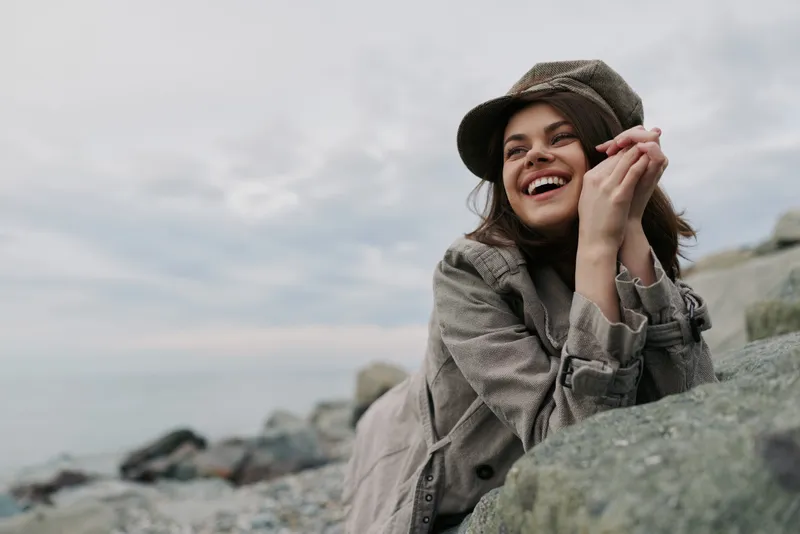 Young woman in stylish coat and cap smiles happily while resting on rocks near the sea shore. Overcast sky and rocky beach create a calm and natural outdoor atmosphere.