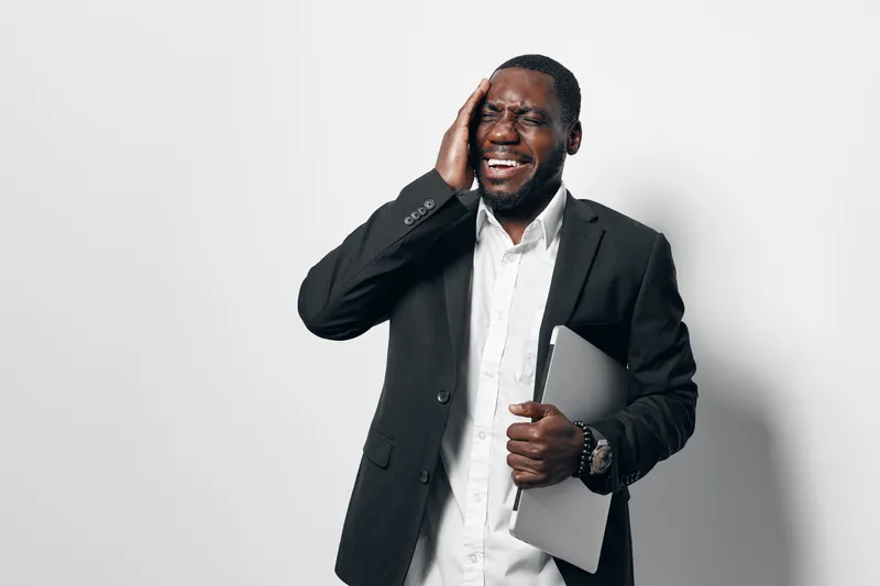 African American man in black suit holding laptop with hand on head showing stress or headache isolated on white background. Businessman, frustration and technology concept.