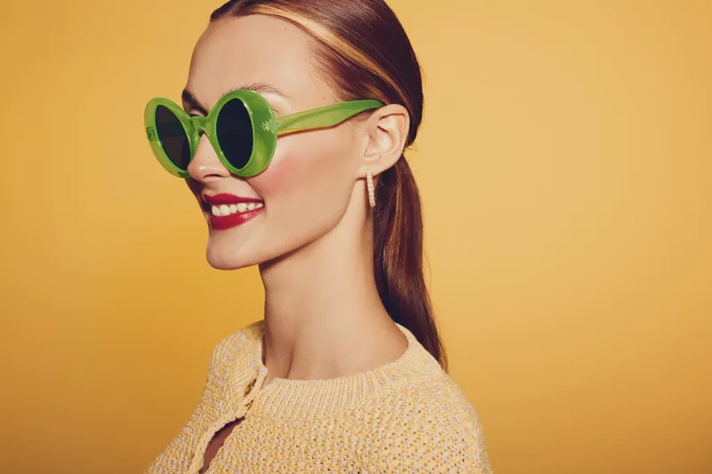 Smiling woman with green round sunglasses and red lipstick poses against yellow background. Stylish female model wears beige sweater and gold earrings, showing confident and happy mood.