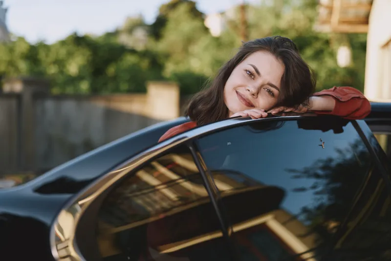 Young woman with brunette hair leans on a sleek car, smiling warmly at the camera, conveying casual confidence and a candid everyday lifestyle moment